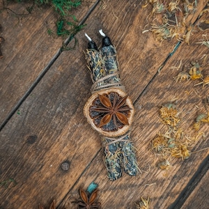 May include: Aromatic bundle with two black candles, dried orange slice, and star anise on a rustic wooden surface. The bundle is wrapped with twine and adorned with dried herbs and flowers, creating a natural, earthy aesthetic.