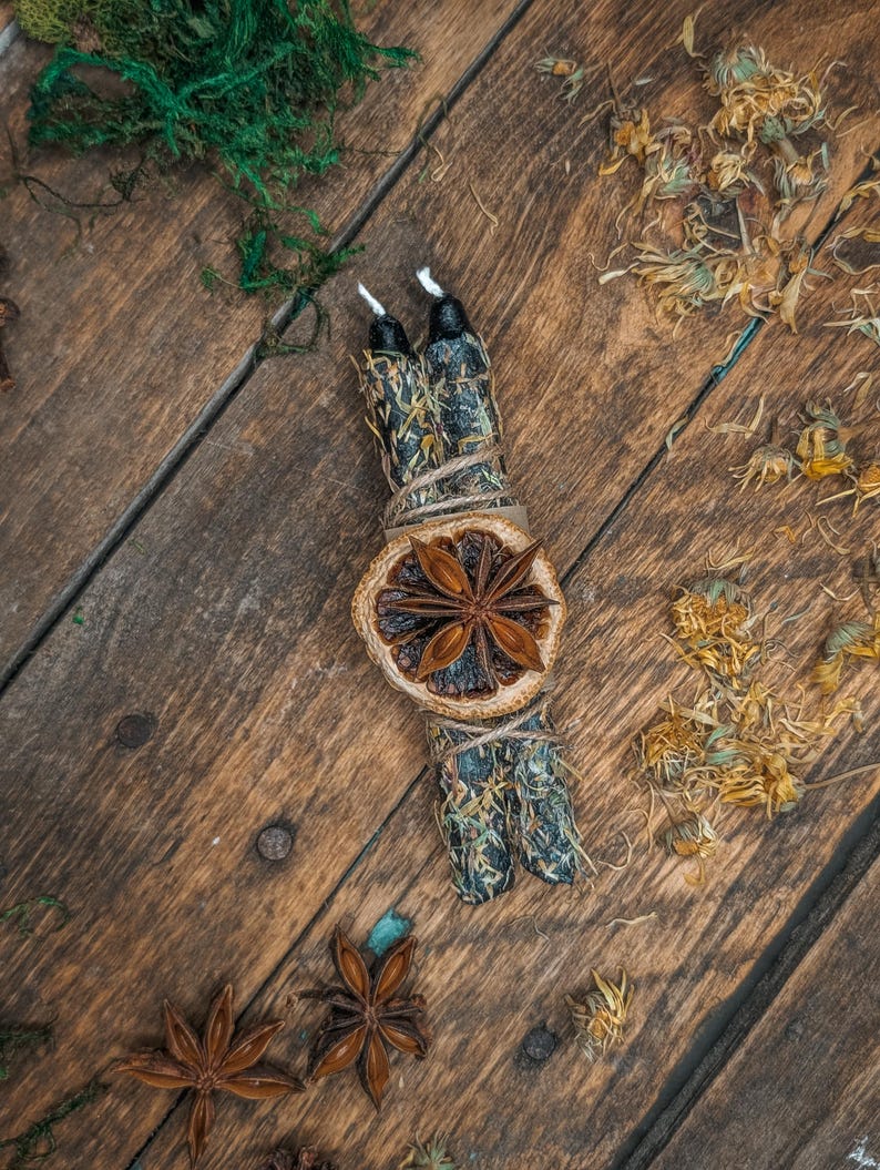 May include: Aromatic bundle with two black candles, dried orange slice, and star anise on a rustic wooden surface. The bundle is wrapped with twine and adorned with dried herbs and flowers, creating a natural, earthy aesthetic.