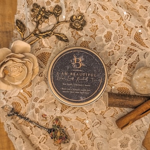 May include: A round tin with the text "I AM BEAUTIFUL Love Look Nailah Tea" sits on a cream lace cloth. The tin is surrounded by a decorative bee branch, a wooden rose, a spoon with dried flowers, and cinnamon sticks.