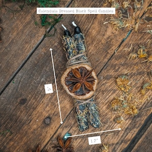 May include: Two black spell candles, 5 inches tall, wrapped with dried herbs and twine, and adorned with a dried orange slice and star anise. The text on the image reads "Calendula Dressed Black Spell Candles".