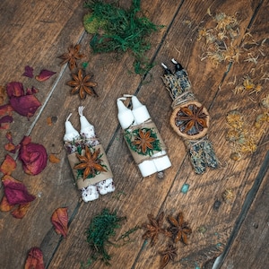 May include: An overhead shot of three sets of white candles, each bundled and decorated with star anise, twine, and greenery. Dried rose petals and star anise are scattered on the wooden surface, creating a rustic, natural aesthetic.