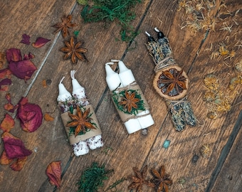 Op de afbeelding: Een bovenaanzicht van drie sets witte kaarsen, elk gebundeld en versierd met steranijs, touw en groen. Gedroogde rozenblaadjes en steranijs zijn verspreid over het houten oppervlak, wat een rustieke, natuurlijke uitstraling creëert.
