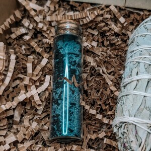 May include: A small glass bottle filled with blue crystals, sitting in brown packing peanuts. A bundle of white sage is visible in the background.