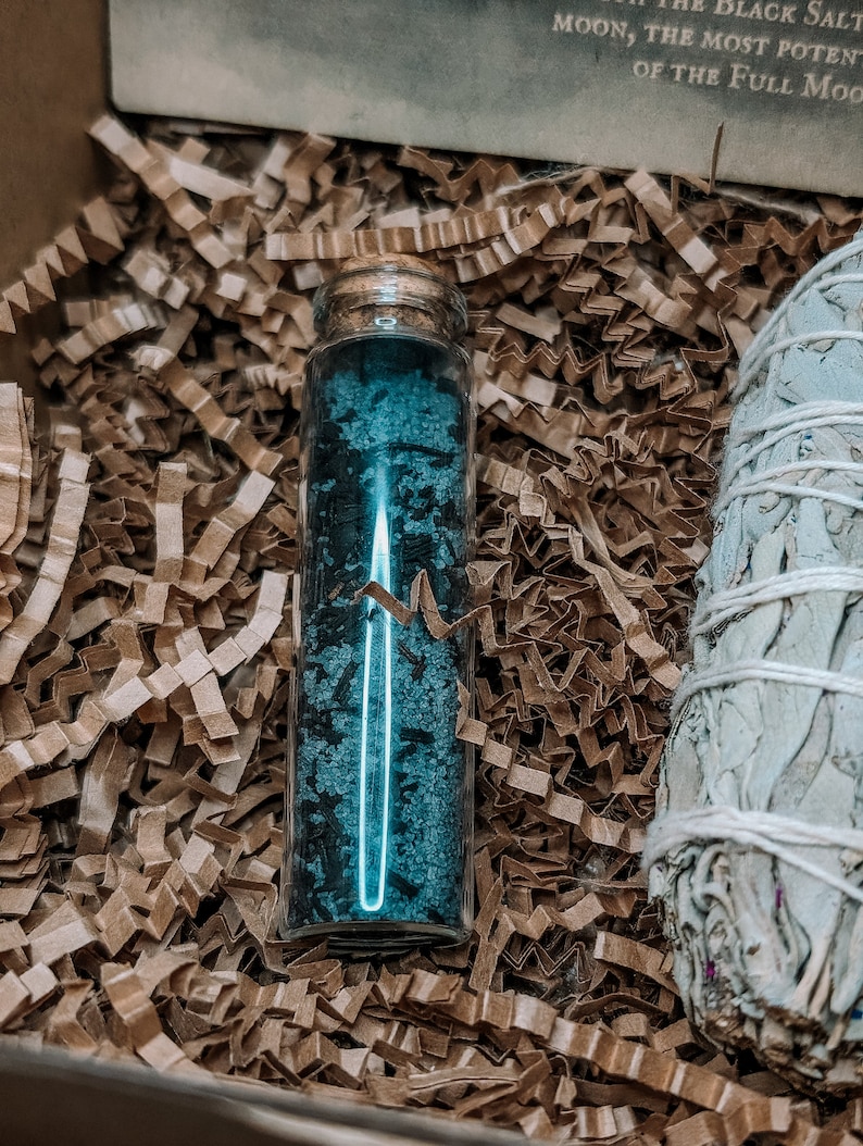 May include: A small glass bottle filled with blue crystals, sitting in brown packing peanuts. A bundle of white sage is visible in the background.