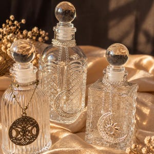 May include: Three clear glass bottles with spherical stoppers, each featuring a decorative metal charm hanging from a chain around the neck. One bottle has a round pendant with a triple moon symbol, while the others feature crescent moon charms.