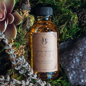 May include: A clear glass bottle with a black cap, filled with amber liquid, and a brown label that reads "New Moon Ritual Oil." The bottle is surrounded by green moss, a succulent, dried lavender, and a crystal.