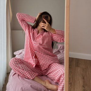 Puede incluir: Un conjunto de pijama de cuadros vichy rosa y blanco con mangas largas y pantalones. La persona que lleva el pijama est&aacute; sentada en una cama con una manta rosa.
