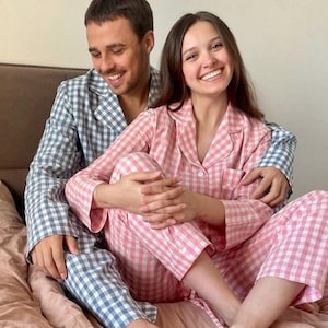 Puede incluir: Una pareja vistiendo pijamas a juego de cuadros azules y rosas sentados en una cama. Sonr&iacute;en y se miran.