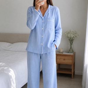 May include: Pale blue pyjama set, comprising a button-up shirt and wide-leg trousers. The set has white trim on the collar, cuffs, and trouser hems. The fabric has a textured finish. The person in the image is holding a phone.