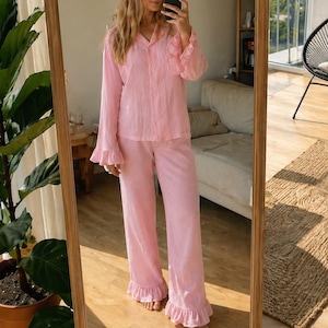 Op de afbeelding: Een lichtroze tweedelige pyjamaset bestaande uit een overhemd met lange mouwen en knoopsluiting en een bijpassende wijde broek. Het overhemd heeft een kraag en ruches aan de mouwen, terwijl de broek is afgewerkt met bijpassende ruches.