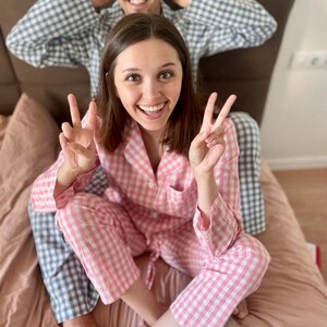Puede incluir: Una pareja lleva pijamas a juego de cuadros rosas y azules. La mujer sonr&iacute;e y hace el signo de la paz con ambas manos. El hombre est&aacute; sentado detr&aacute;s de ella con las manos detr&aacute;s de la cabeza.