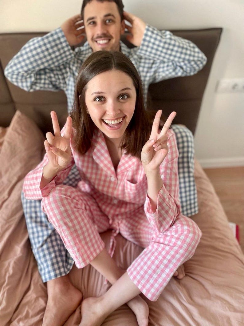 Puede incluir: Una pareja lleva pijamas a juego de cuadros rosas y azules. La mujer sonr&iacute;e y hace el signo de la paz con ambas manos. El hombre est&aacute; sentado detr&aacute;s de ella con las manos detr&aacute;s de la cabeza.