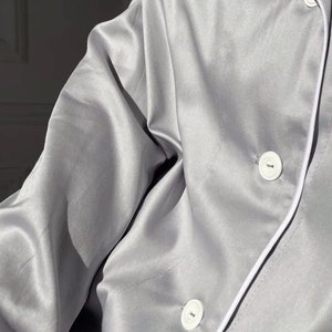 K&ouml;nnte beinhalten: Ein graues Satin-Pyjama-Oberteil mit wei&szlig;en Paspeln und wei&szlig;en Kn&ouml;pfen. Das Oberteil hat eine lockere Passform und einen Kragen.