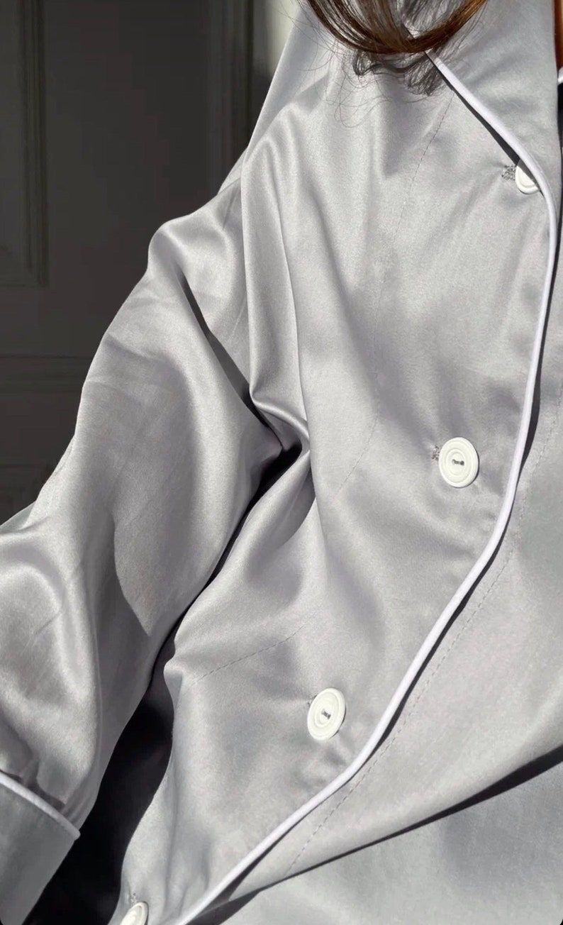 K&ouml;nnte beinhalten: Ein graues Satin-Pyjama-Oberteil mit wei&szlig;en Paspeln und wei&szlig;en Kn&ouml;pfen. Das Oberteil hat eine lockere Passform und einen Kragen.