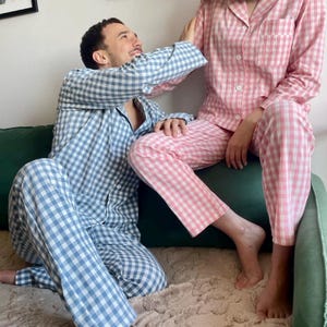 Puede incluir: Una pareja vistiendo pijamas a cuadros a juego. La mujer lleva un pijama rosa y blanco, y el hombre lleva un pijama azul y blanco. Est&aacute;n sentados en un sof&aacute; verde y se sonr&iacute;en.