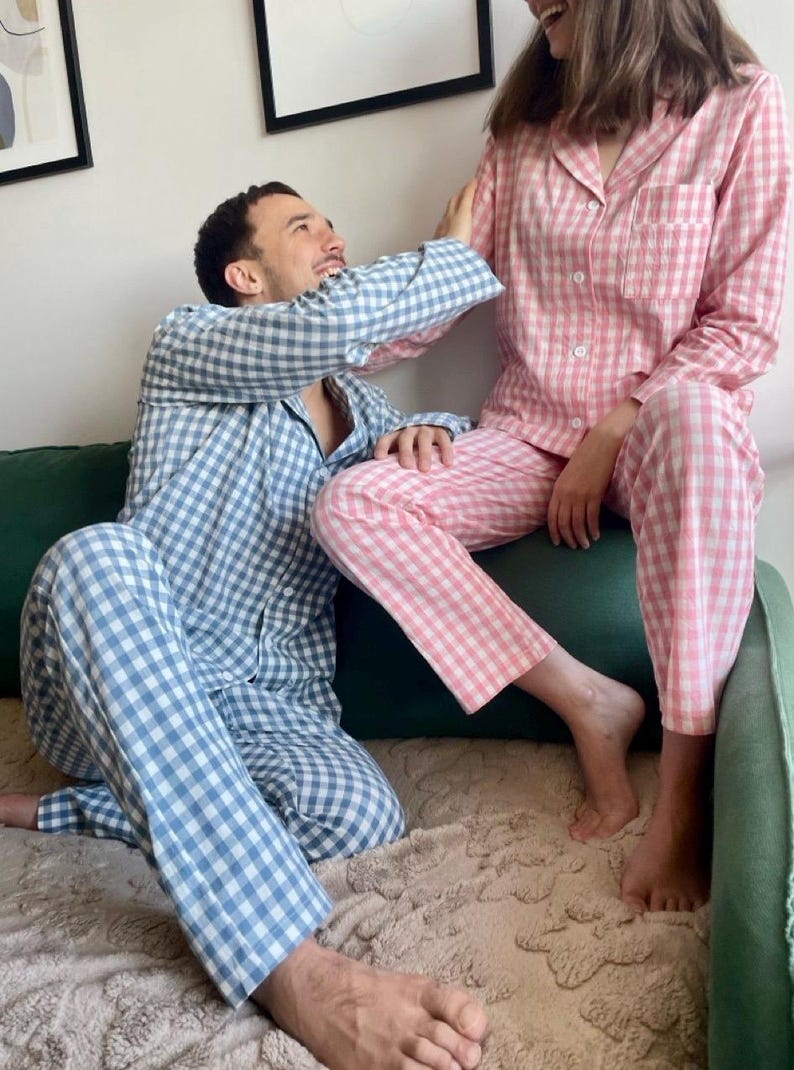 Puede incluir: Una pareja vistiendo pijamas a cuadros a juego. La mujer lleva un pijama rosa y blanco, y el hombre lleva un pijama azul y blanco. Est&aacute;n sentados en un sof&aacute; verde y se sonr&iacute;en.