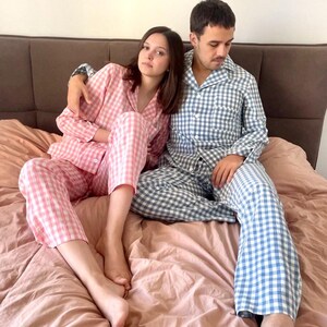 Puede incluir: Una pareja vistiendo pijamas a juego. La mujer lleva un pijama de cuadros rosa y blanco y el hombre lleva un pijama de cuadros azul y blanco. Est&aacute;n sentados en una cama con una manta rosa.