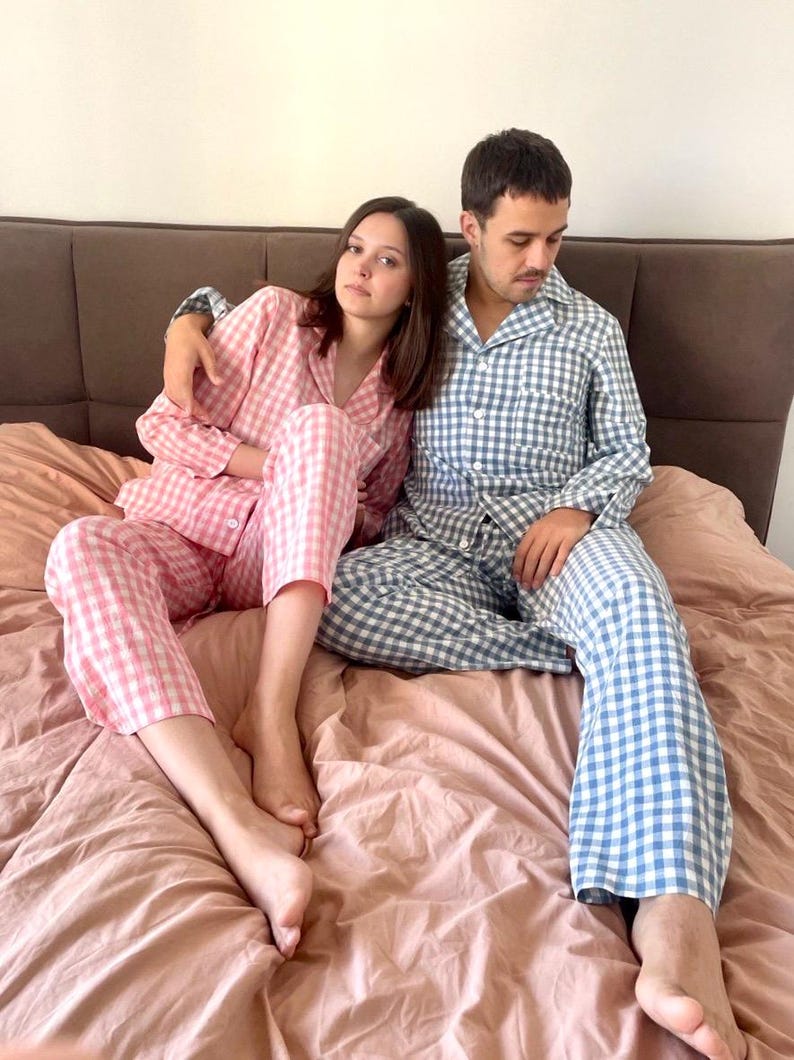 Puede incluir: Una pareja vistiendo pijamas a juego. La mujer lleva un pijama de cuadros rosa y blanco y el hombre lleva un pijama de cuadros azul y blanco. Est&aacute;n sentados en una cama con una manta rosa.