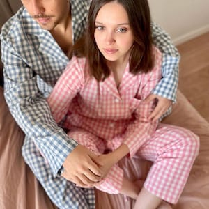 Puede incluir: Una pareja vistiendo pijamas a juego de cuadros azules y rosas. El hombre lleva una camisa de cuadros azules y blancos y la mujer lleva una camisa de cuadros rosas y blancos. Est&aacute;n sentados en una cama con una manta rosa.