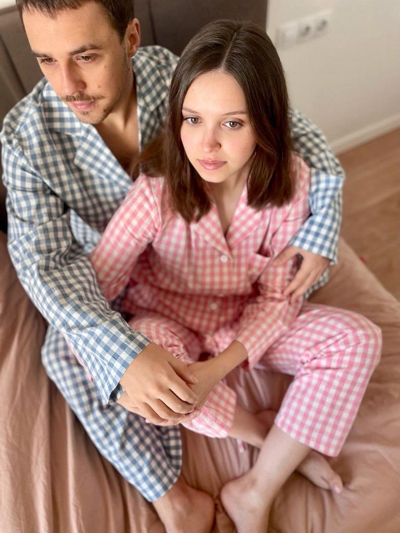 Puede incluir: Una pareja vistiendo pijamas a juego de cuadros azules y rosas. El hombre lleva una camisa de cuadros azules y blancos y la mujer lleva una camisa de cuadros rosas y blancos. Est&aacute;n sentados en una cama con una manta rosa.