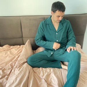 K&ouml;nnte beinhalten: Eine Person tr&auml;gt einen gr&uuml;nen Waffelpiqu&eacute;-Pyjama mit einem Hemd mit Kn&ouml;pfen und einer langen Hose. Die Person sitzt auf einem Bett mit einem braunen Kopfteil und einer rosa Bettdecke.