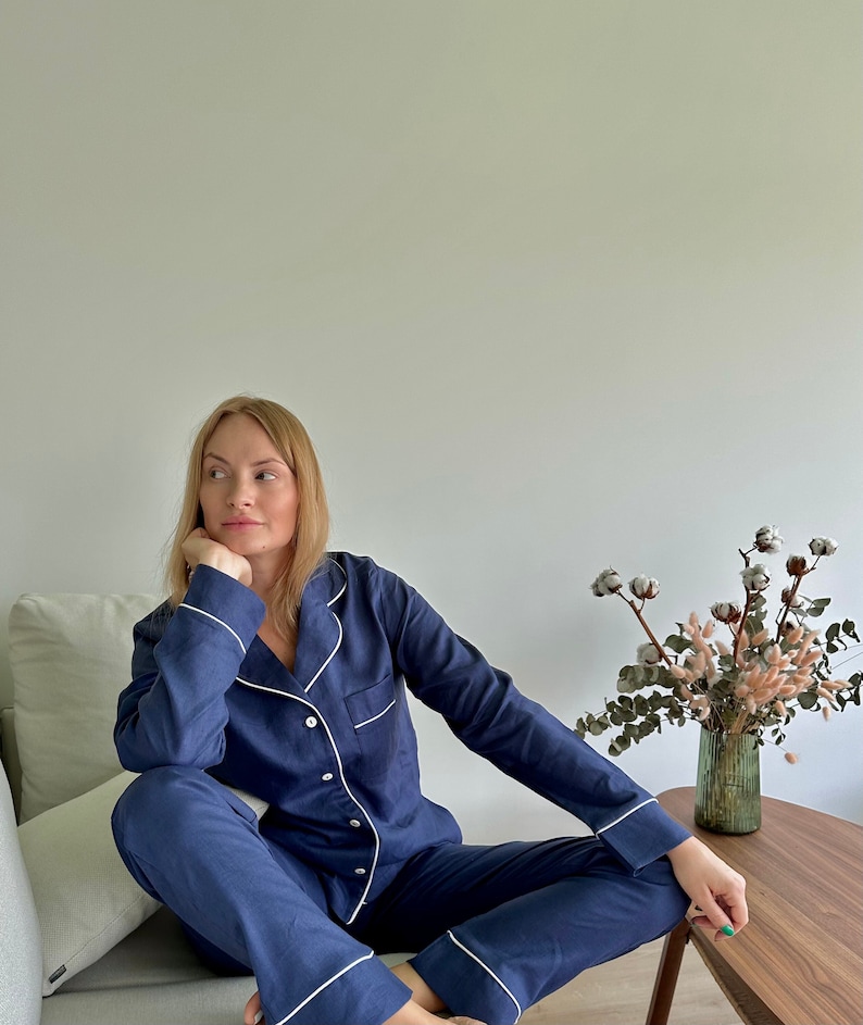 May include: A person wearing a navy blue pajama set with white piping. The pajamas have a button-up top and long pants. The person is sitting on a couch with a white throw pillow.