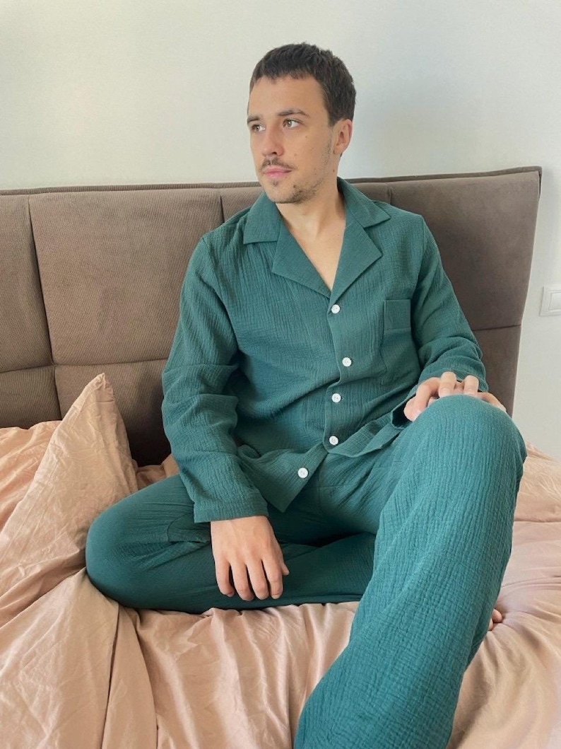 K&ouml;nnte beinhalten: Eine Person tr&auml;gt einen gr&uuml;nen Schlafanzug mit einem Hemd mit Kragen und einer langen Hose. Die Person sitzt auf einem Bett mit einer rosa Decke.