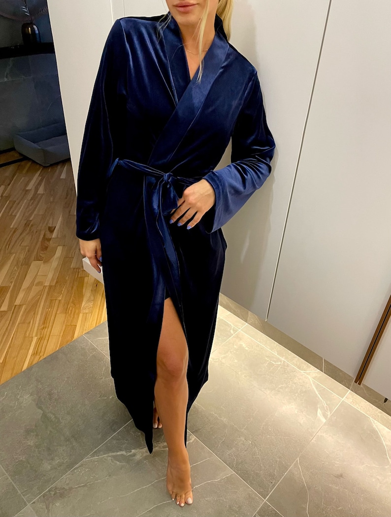 May include: A long, navy blue velvet robe with a shawl collar and a tie belt. The robe has a slit up the front and long sleeves.