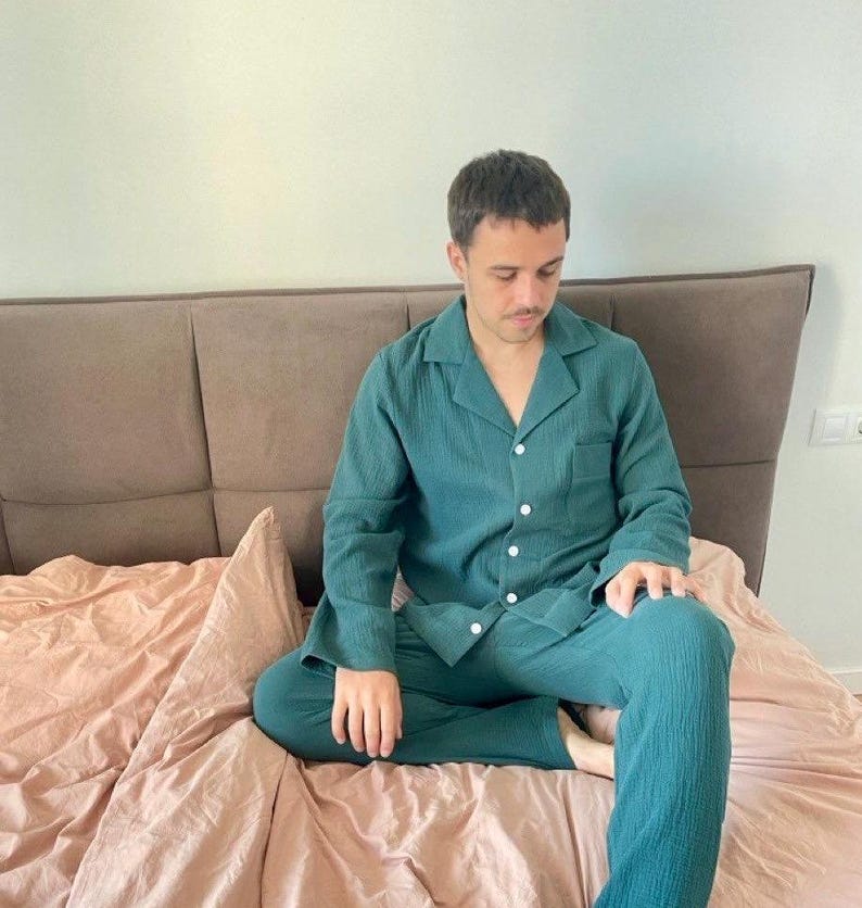 K&ouml;nnte beinhalten: Eine Person tr&auml;gt ein dunkelgr&uuml;nes, lang&auml;rmliges Pyjama-Oberteil und eine passende Hose. Der Pyjama hat einen Knopfleisten vorne und eine lockere Passform. Die Person sitzt auf einem Bett mit einer hellrosa Bettdecke.