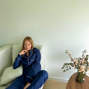 May include: A person wearing a navy blue pajama set with white piping. The pajamas have a button-up top and long pants. The person is sitting on a white couch.