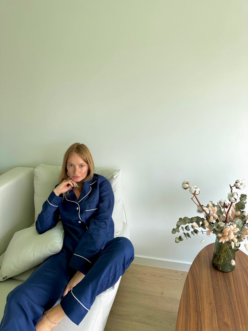 May include: A person wearing a navy blue pajama set with white piping. The pajamas have a button-up top and long pants. The person is sitting on a white couch.