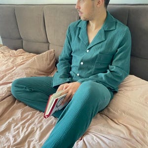 K&ouml;nnte beinhalten: Eine Person tr&auml;gt einen tealblauen Schlafanzug mit einem lang&auml;rmeligen Hemd und einer Hose. Die Person sitzt auf einem Bett und h&auml;lt ein Buch.