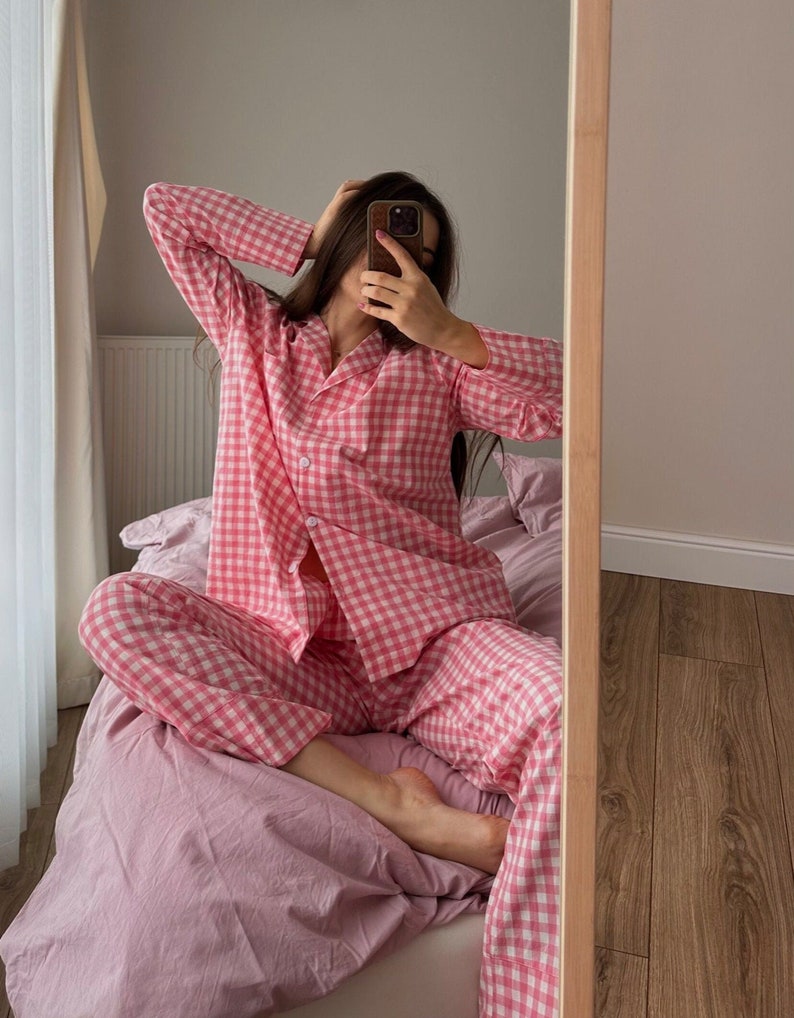 Puede incluir: Un conjunto de pijama de cuadros vichy rosa y blanco con mangas largas y pantalones. La persona que lleva el pijama est&aacute; sentada en una cama con una manta rosa.