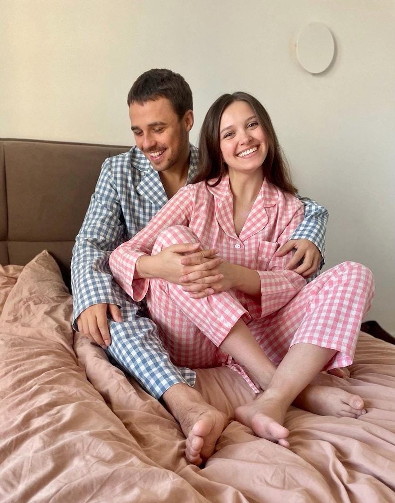 Puede incluir: Una pareja vistiendo pijamas a juego de cuadros azules y rosas sentados en una cama. Sonr&iacute;en y se miran.