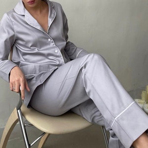 K&ouml;nnte beinhalten: Ein hellgrauer Satin-Pyjama mit wei&szlig;en Paspeln. Das Oberteil hat einen Kragen und lange &Auml;rmel. Die Hose hat eine gerade Bein und einen wei&szlig;en Saum an den B&uuml;ndchen.