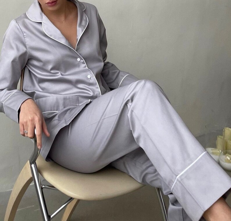 K&ouml;nnte beinhalten: Ein hellgrauer Satin-Pyjama mit wei&szlig;en Paspeln. Das Oberteil hat einen Kragen und lange &Auml;rmel. Die Hose hat eine gerade Bein und einen wei&szlig;en Saum an den B&uuml;ndchen.