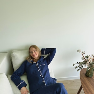 May include: A woman wearing a navy blue pajama set with white piping. She is sitting on a couch with a white pillow and looking to the side.