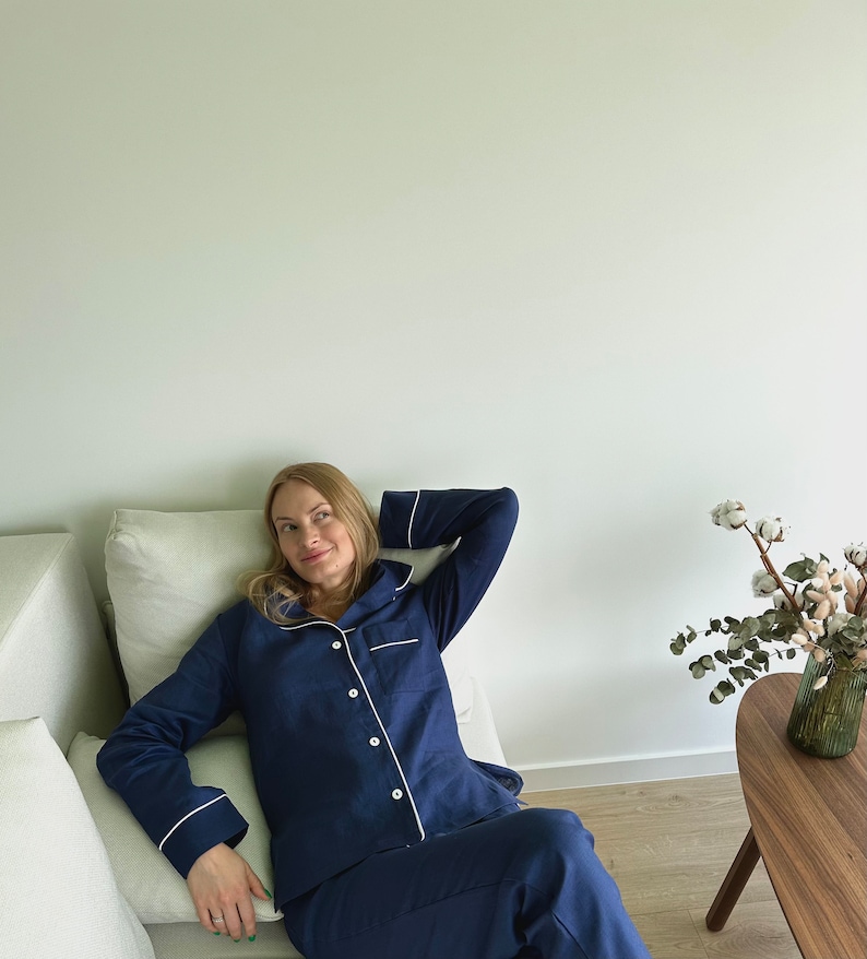 May include: A woman wearing a navy blue pajama set with white piping. She is sitting on a couch with a white pillow and looking to the side.
