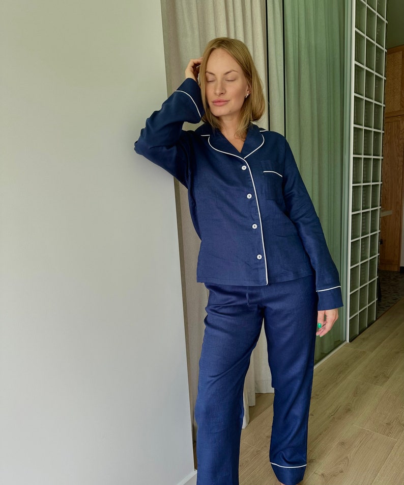 May include: A navy blue pajama set with white piping. The top has a long sleeve and a button-up front. The pants are long and have a straight leg.