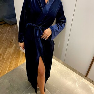 May include: A long, navy blue velvet robe with a shawl collar and a tie belt. The robe has a slit up the front and long sleeves.