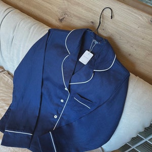 May include: A navy blue long-sleeved pajama top with white piping around the collar, cuffs, and pockets. The top has a button closure and a small white tag with the text "GOODNIGHT" on it.