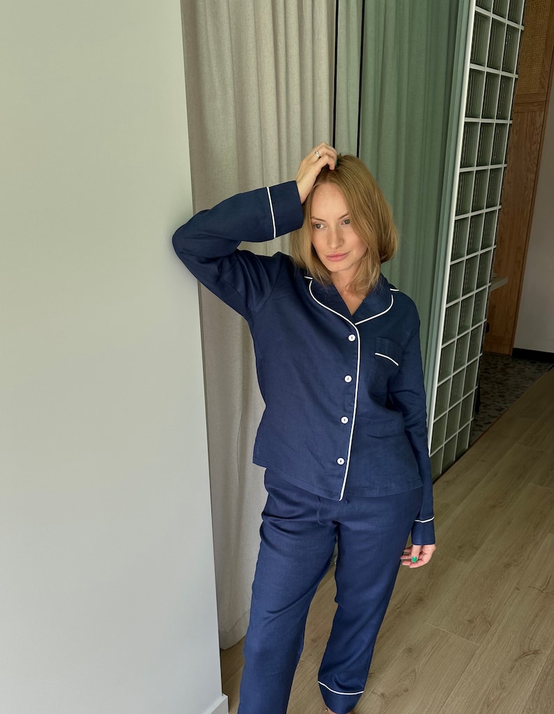 May include: A navy blue pajama set with white piping. The top has a button-down front and long sleeves. The pants have a straight leg and a white trim at the bottom.