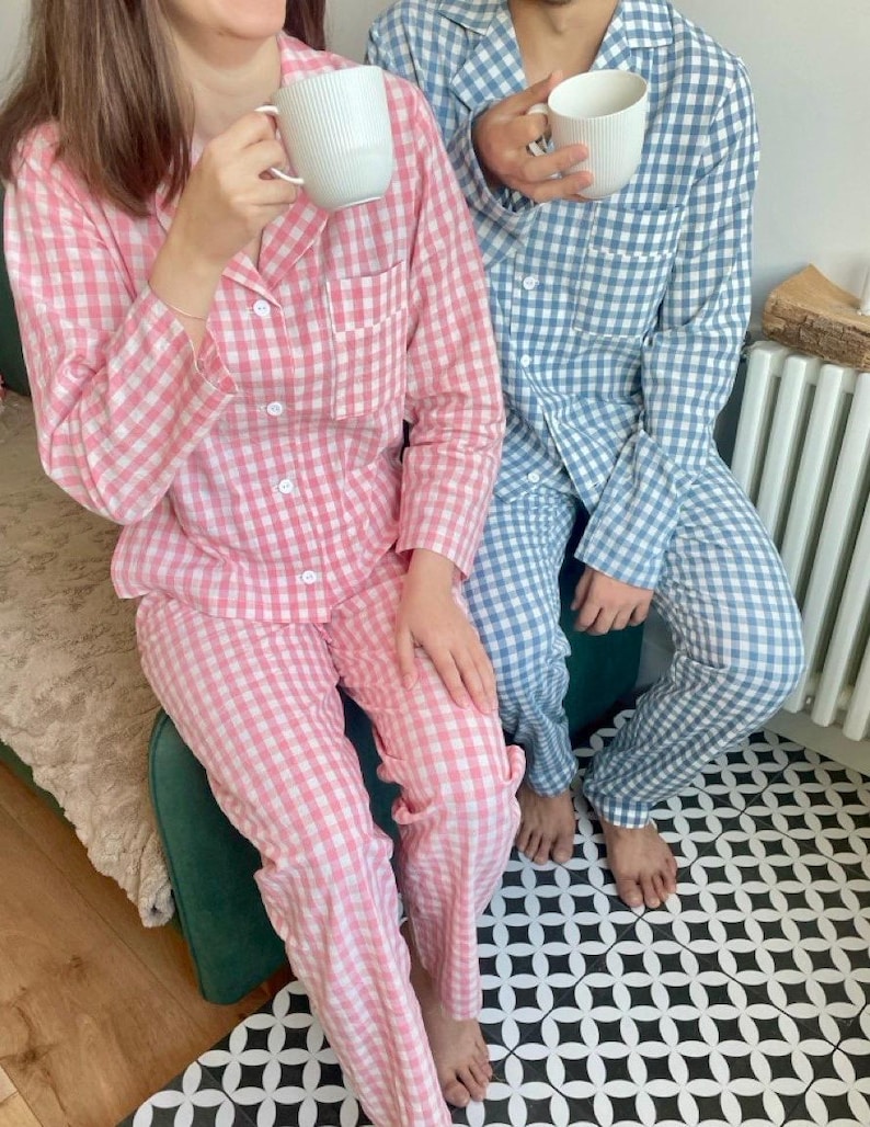 Puede incluir: Dos personas vistiendo pijamas a cuadros a juego. Una persona lleva un pijama a cuadros rosa y blanco y la otra lleva un pijama a cuadros azul y blanco. Ambas personas sostienen tazas blancas.