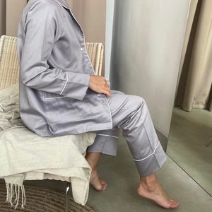 K&ouml;nnte beinhalten: Ein grauer Satin-Pyjama mit wei&szlig;en Paspeln. Das Oberteil hat lange &Auml;rmel und einen Knopfleisten vorne. Die Hose hat eine gerade Beinpartie.