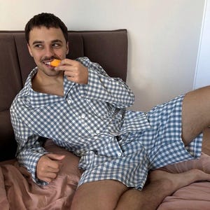 Puede incluir: Una persona vistiendo un pijama a cuadros azul y blanco. La parte superior del pijama tiene cuello y mangas largas. La parte inferior del pijama es un short. La persona est&aacute; sentada en una cama y comiendo una pieza de fruta.