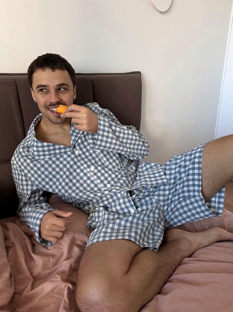 Puede incluir: Una persona vistiendo un pijama a cuadros azul y blanco. La parte superior del pijama tiene cuello y mangas largas. La parte inferior del pijama es un short. La persona est&aacute; sentada en una cama y comiendo una pieza de fruta.