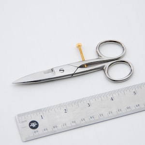 Può includere: Un paio di piccole forbici chirurgiche argentate con una vite dorata. Le forbici sono lunghe circa 10 cm e sono su una superficie bianca accanto a un righello.