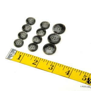 May include: A set of 10 black and white buttons with four holes each. The buttons are arranged in three rows on a white surface. A yellow measuring tape is in the lower left corner of the image.