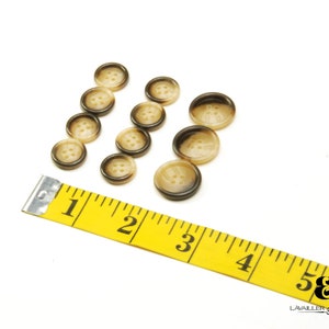 May include: A set of ten brown and beige buttons with four holes each. The buttons are arranged in a semi-circle shape on a white surface. A yellow measuring tape is in the lower right corner of the image.