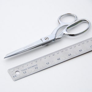May include: A silver pair of scissors with a ruler measuring 20 inches. The scissors have a curved handle and a sharp blade.